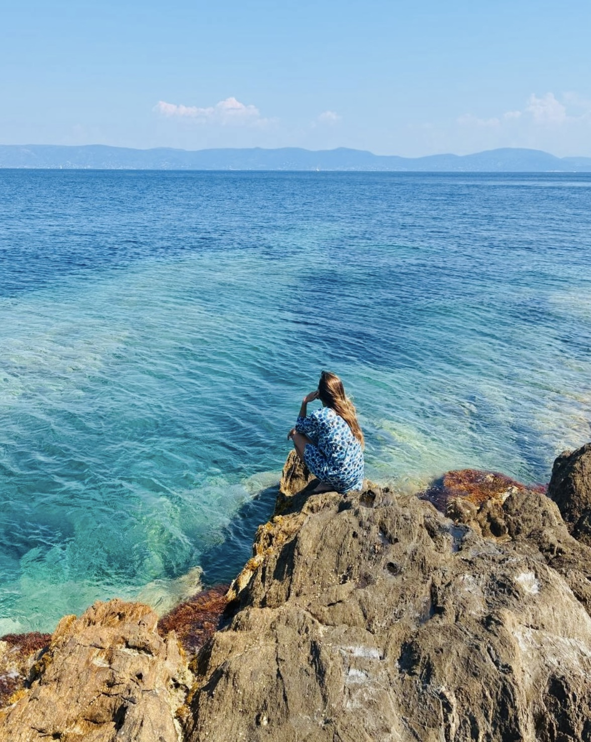 Isa, robe bleue, accroupie sur rocher face océan